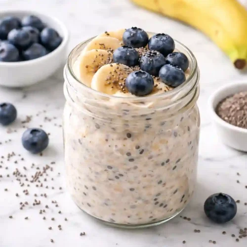 Frasco de vidrio lleno de avena nocturna cremosa con arándanos frescos y rodajas de plátano, sobre superficie de mármol blanco con semillas de chía esparcidas