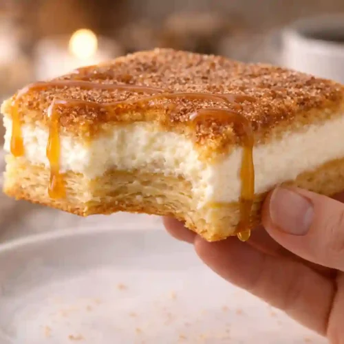 Sosteniendo una barra de cheesecake de churros con mordida que revela el relleno cremoso y la cobertura de canela